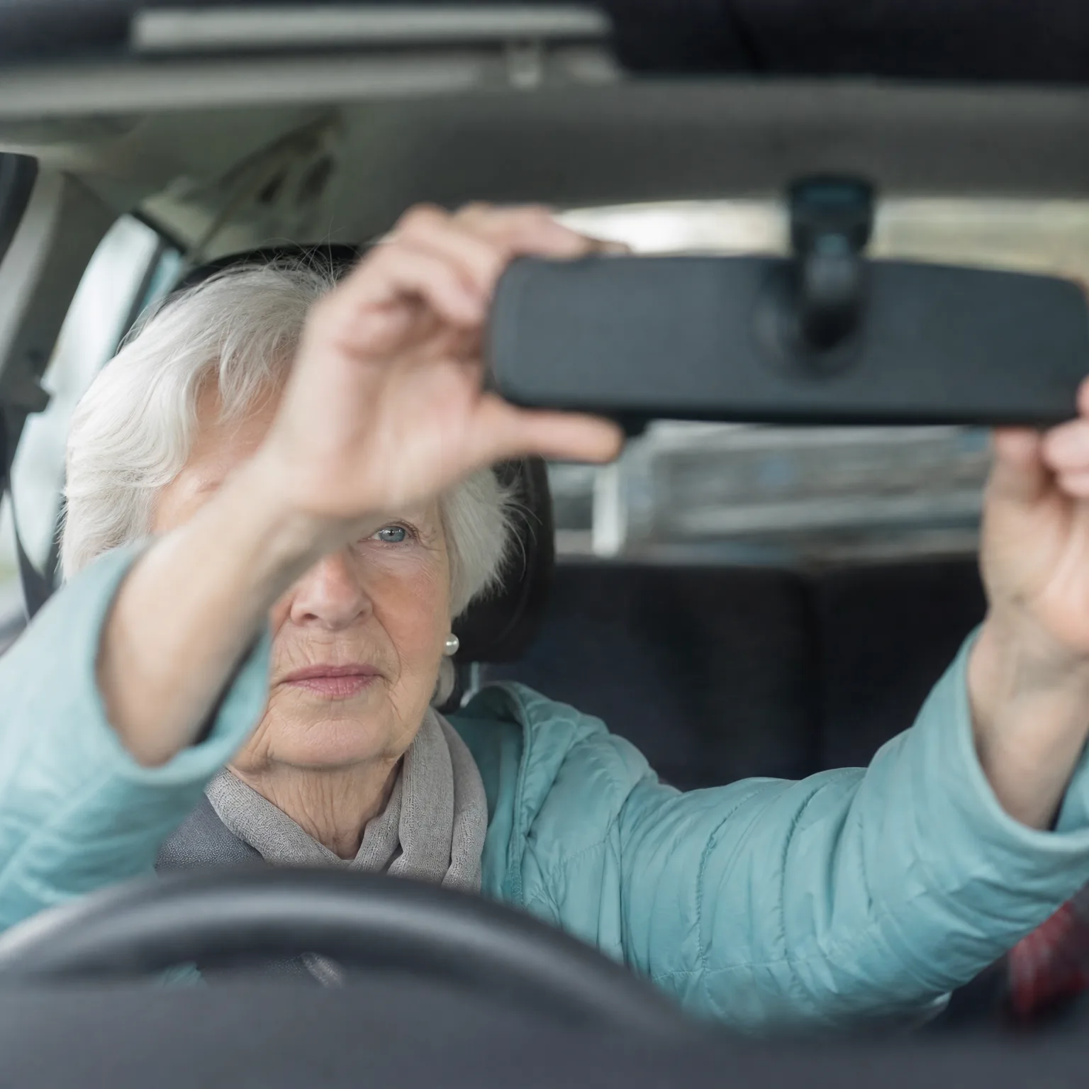 An older woman with short white hair in a light teal jacket sits in a car's driver seat and reaches up with both hands to adjust the rear-view mirror.